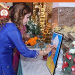 Woman in a blue kurta performs a ritual at a shrine, touching a framed portrait of Sai Baba amid marigolds and decorative gifts.