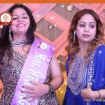 Two women in ornate Indian outfits at a festive event; one wears a lavender sash while smiling, the other looks aside on a decorated stage.