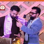 Two men at a celebration; one wearing a purple sash that reads 'MR HERITAGE' receives a microphone from the other man, beside a wrapped gift.