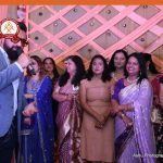 Man in a blue blazer sings into a microphone while a line of women in colorful sarees watches at a festive event, logo visible in the top-left corner.