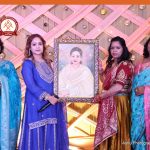 Four women in colorful traditional outfits pose with a framed portrait of a woman on a decorative stage backdrop.