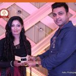 Man presenting an award to a smiling woman at a decorated event backdrop, logo top left.
