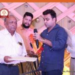 Man in a blue shirt hands a microphone and an orange cloth to an elderly man in a striped shirt during an awards ceremony, with two onlookers and a pink-gold decorative backdrop.