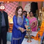 Woman in a blue traditional outfit lights a ceremonial lamp during a festive event, with onlookers and marigold garlands in the backdrop.