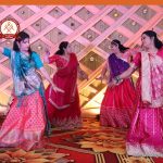 Group of women in bright sarees dancing on a stage with a geometric lattice backdrop and warm lighting.