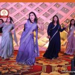 Five women in colorful sarees dancing on a decorated stage with a pink lattice backdrop and a photographer watermark in the corner.