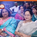 Two women in colorful sarees sit on a blue couch at a formal event, with attendees and a logo visible in the background.