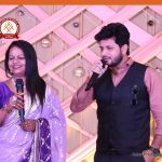 Woman in a purple sari and a man in a pink shirt with a black vest sing into handheld microphones on a decorated stage.