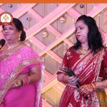 Two Indian women in colorful sarees stand on stage, each holding a microphone, as they perform against a decorative pink backdrop with lights and a logo in the corner.