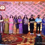 Line of eleven people in colorful traditional outfits stand in a row on a decorated stage, each holding a wrapped gift, with a floral lattice backdrop and stage lights.
