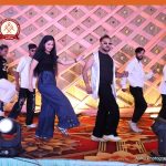 Group of people dancing on a decorated stage with colorful lighting and floral carpet pattern, mid-performance.