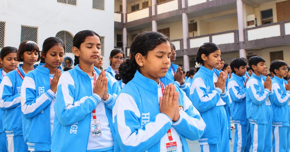 Group of students in blue tracksuits standing in a school courtyard with hands in prayer during a morning assembly (formation, calm, focused).