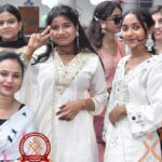 Group of young women in white traditional outfits posing for a photo at a school event, with balloons in the background and the Heritage International School logo visible.