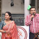Man in a red checkered shirt speaks into a microphone beside a woman in a red sari at a school event with a circular school banner in the foreground.