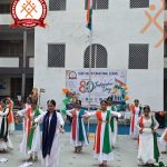 Group of girls in white dresses with orange, white, and green sashes performing a dance on a school courtyard during Independence Day celebrations in front of a banner and Indian flag.